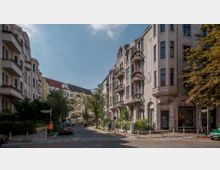 Straßenszene in einem Wohngebiet mit mehrstöckigen Altbauhäusern im Gründerzeitstil, großen Fenstern, Erkern und Balkonen. Der Straßenverlauf ist gesäumt von Bäumen, parkenden Autos und Verkehrsschildern, im Hintergrund sind weitere Gebäude sichtbar.