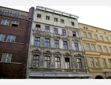 Eine Häuserreihe mit drei nebeneinanderliegenden Gebäuden, darunter ein rotes Klinkergebäude, ein sanierungsbedürftiges, graues Gebäude mit dekorativen Fensterrahmen und Verzierungen sowie ein renoviertes, gelbes Gebäude mit symmetrischen Fenstern.