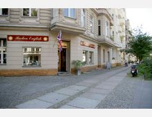 Eckgebäude mit hellem Putz und einer Ladenfront, die mit einem roten Schild "Broken English" und einer britischen Flagge dekoriert ist; davor ein Bürgersteig mit Kopfsteinpflaster und ein Gehweg, umgeben von weiteren Wohnhäusern und Bäumen.