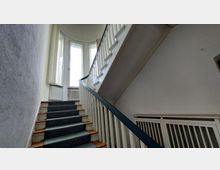 Treppenhaus mit einer hölzernen Treppe, die links eine Biegung macht. Die Treppe hat ein dunkel lackiertes Geländer und führt zu einem Bereich mit hohen, halbkreisförmigen Fenstern, die viel Tageslicht einlassen.