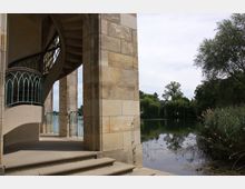 Steinbogenkonstruktion mit einer Wendeltreppe und grünem Geländer, daneben ein See mit Schilf, Bäumen und ruhiger Wasserfläche im Hintergrund.