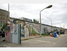 Teil der Berliner Mauer mit farbenfrohen Graffiti, darunter das bekannte Motiv des „Bruderkusses“. Der Bereich um die Mauer zeigt Mülleimer, Fahrräder und eine offene Metalltür, während im Hintergrund moderne und historische Gebäude zu sehen sind.