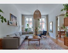 Helles Wohnzimmer mit einer grauen Eckcouch, einem Holztisch und einem blauen Schaukelstuhl. Große Fenster mit Vorhängen, zahlreiche Zimmerpflanzen und eine hängende Rattanleuchte prägen den Raum; durch eine Tür ist ein Esszimmer sichtbar.