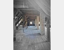 Dachboden mit Holzkonstruktion und Holzboden, unterstützt von sichtbaren Balken und Säulen. Am Ende des Raumes sind zwei Fenster erkennbar, die Tageslicht hereinlassen.