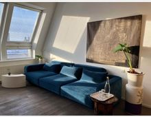 Modern living room with a large blue sectional sofa beneath a slanted ceiling and tall window with blinds casting light patterns on white walls. An abstract wall artwork, small round tables, and potted plants add decor on a dark wood floor.