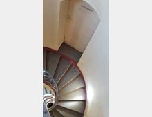 Eine spiralförmige Treppe mit Metallgeländer, die nach unten führt, in einem hellen Innenraum mit beige gestrichenen Wänden. Über dem unteren Treppenabsatz ist der Schriftzug „Amtsgericht“ an der Wand zu sehen.