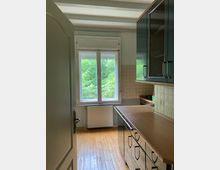 Eine kleine Küche mit Holzboden, einem großen Fenster mit Blick ins Grüne und einer gefliesten Rückwand. Rechts befinden sich schwarze Schränke mit glänzenden Oberflächen und eingebauten Geräten.