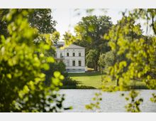 Weißes zweigeschossiges Herrenhaus mit symmetrischer Fassade, umgeben von einer Parklandschaft mit großen Bäumen und einer Wiese, im Vordergrund ein See mit Uferbewuchs.
