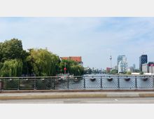 Brücke mit dekorativem Geländer, im Hintergrund ein Fluss gesäumt von Bäumen und modernen Gebäuden. Der Berliner Fernsehturm und andere Bauwerke ragen in die skyline, bei klarem Himmel im Hintergrund.