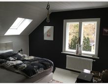 Schlafzimmer in einem Dachgeschoss mit weißer Holzdecke und einer Dachschräge mit Dachfenster. Ein Doppelbett mit dunkler, floraler Bettwäsche steht vor einer schwarz gestrichenen Wand, daneben ein Fenster mit Blick auf Bäume.