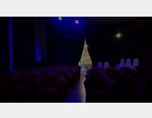 Dunkler Raum mit mehreren Reihen roter Polstersessel, die nach vorne ausgerichtet sind; im Hintergrund ein durch Vorhänge abgetrennter Bereich mit heller Beleuchtung, in dem weiße Stühle und ein Tisch sichtbar sind.