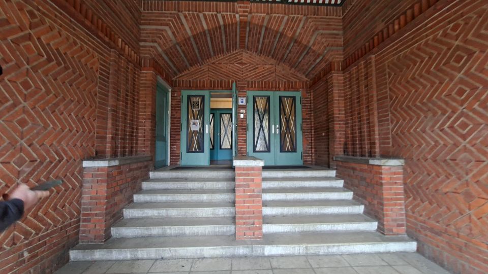 Überdachter Eingangsbereich eines Backsteingebäudes mit gemustertem Ziegelgewölbe, breiter Treppe und mittigem Ziegelpfeiler. Oben befinden sich grün gestrichene Doppeltüren mit verglasten Einsätzen und Rautengittern.
