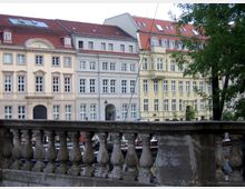 Reihe von historischen Wohngebäuden mit hellen Fassaden und roten Dächern, aufgenommen hinter einem steinernen Geländer; im Vordergrund ein kleiner Wasserkanal mit Booten und angrenzenden Bäumen.