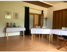 Ein heller Raum mit Holzboden, in dem mehrere Tische mit weißen Tischdecken stehen. An der Wand befinden sich zwei Schmetterlingsbilder, daneben ein gedeckter Tisch mit Wasserflaschen, Gläsern und einem Obstständer. Im Hintergrund ist eine geöffnete Tür zu sehen, die in einen angrenzenden Raum führt.