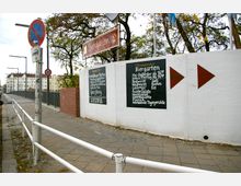 Eine Biergartenanlage mit weißen Wänden, auf denen Schilder mit Öffnungszeiten und Angeboten wie Speisen und Getränke angebracht sind. Im Vordergrund sind ein Gehweg, ein Verkehrsschild und ein Zaun zu sehen, im Hintergrund befinden sich Bäume und mehrstöckige Wohnhäuser.