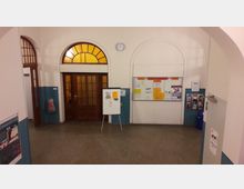 Ein Flur mit einer Tür aus Holz und Glas in der Mitte, flankiert von jeweils einer weiteren Glastür. Darüber befindet sich ein halbkreisförmiges Fenster mit gelbem Glas. An der rechten Wand hängt ein großes schwarzes Brett mit Notizen und Informationen, daneben Prospekthalter und ein blauer Mülleimer. In der Mitte des Raums steht eine Stellwand mit weiteren Informationen.