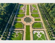 Symmetrischer Barockgarten mit geometrisch angelegten Rasenflächen, symmetrischen Blumenmustern und einem achteckigen Wasserbecken in der Mitte, umgeben von Kieswegen und Bäumen. Der Garten befindet sich in einer waldreichen Umgebung.