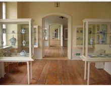 Museumsausstellungsraum mit Holzfußboden, gläsernen Vitrinen und ausgestelltem blau-weißem Porzellan. Durch eine Reihe offener Türen sind weitere Räume zu sehen.