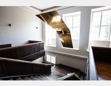 Holztreppe in einem historischen Gebäude mit dunklem Holzgeländer, großen Fenstern, die Tageslicht hereinlassen, und einer schwebenden, goldenen Metallinstallation im Treppenbereich.