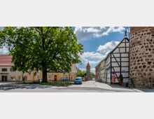 Historischer Dorfplatz mit Fachwerkhäusern, einem großen Baum in der Mitte und einer gepflasterten Straße, die zu einem Kirchturm im Hintergrund führt; rechts ein Teil einer alten Steinmauer.