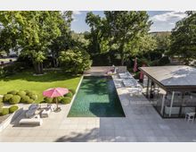 Moderner Garten mit rechteckigem Swimmingpool, umgeben von einer großzügigen Terrasse mit Lounge-Möbeln und Sonnenschirmen. Im Hintergrund eine gepflegte Rasenfläche mit Bäumen und Sträuchern, rechts ein modernes Gebäude mit großen Glasfronten.