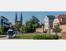 Straßenszene mit einem geparkten Reisebus im Vordergrund, einem historischen Ziegelgebäude und einer Grünfläche. Im Hintergrund ragen die spitzen Türme einer gotischen Kirche über die umliegenden Bäume und Gebäude hervor.