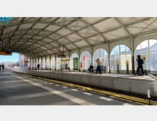 Überdachter, lichtdurchfluteter Bahnsteig einer S-Bahn-Station mit großen bogenförmigen Fenstern, mehreren Personen, die auf den Zug warten, sowie Anzeigetafeln und Sitzbänken auf dem Bahnsteig. Im Hintergrund sind Gebäude und Bäume sichtbar.