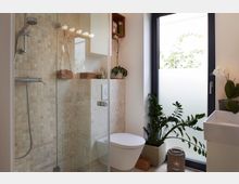 Modernes Badezimmer mit beigefarbenen Mosaikfliesen, einer verglasten Duschkabine, einem hängenden WC und einem großen Fenster mit satiniertem Glas. Pflanzen und dekorative Körbe sorgen für eine natürliche Atmosphäre.