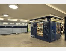 U-Bahn-Station mit einer mit blauen Fliesen verkleideten Wand, auf der "Frankfurter Tor" steht, sowie einem kleinen Kiosk, der Zeitungen und Tabakwaren verkauft. Der Kiosk hat blau gestrichene Wände, die mit Graffiti bedeckt sind, und beleuchtete Regale mit Waren.