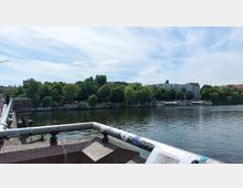 Blick von einer Brücke auf einen Fluss, umgeben von dichter grüner Vegetation und Gebäuden im Hintergrund. Am Ufer sind Weidenbäume, Graffiti und ein Bereich mit Sitzgelegenheiten und Sonnenschirmen zu sehen.