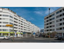 Straßenszene in einem städtischen Viertel mit modernen, weißen Wohngebäuden, die mehrere Stockwerke und Balkone haben. Im Vordergrund verläuft eine Kreuzung mit parkenden und fahrenden Autos, Straßenschildern (eins mit der Aufschrift "Charlottenburg") sowie Gehwegen.