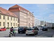 Straßenszene mit mehreren Fahrzeugen, darunter Autos, ein Motorrad und ein Lkw, vor historischen Gebäuden mit roten Ziegeldächern und beige gefärbten Fassaden; im Hintergrund moderne Bürogebäude. Klarer Himmel und tagsüber helle Beleuchtung.
