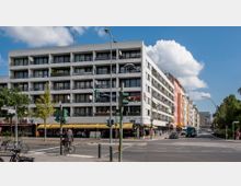 Straßenszene mit einem fünfstöckigen Wohn- und Geschäftsgebäude mit Balkonen und Geschäften im Erdgeschoss, darunter ein Laden mit gelbem Markisendach. Mehrere Verkehrslampen, Fahrräder und Autos sind auf der Straße, während Fußgänger die Kreuzung überqueren; im Hintergrund weitere Gebäude und Bäume.