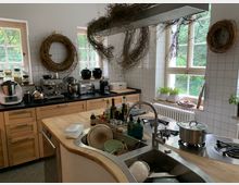 Geräumige, helle Küche mit Holzschränken, einer Arbeitsplatte aus schwarzem Stein und modernen Küchengeräten wie Kaffeemaschine und Mixer. Weiße Fliesenwände, drei Fenster mit Topfpflanzen auf der Fensterbank und dekorative Zweigkränze an den Wänden; das Kochfeld und die Spüle sind in einer abgerundeten Holzinsel integriert.