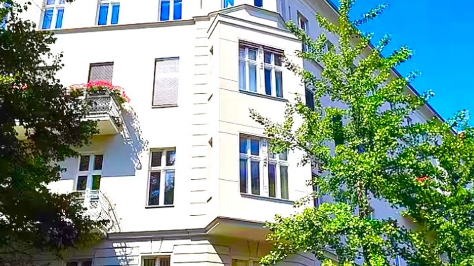 Mehrstöckiges, hell verputztes Eckwohnhaus im Gründerzeitstil mit hohen Fenstern, Erkern und einem kleinen Balkon, aufgenommen aus Straßenperspektive. Im Vordergrund stehen grüne Straßenbäume, darüber ein klarer blauer Himmel.