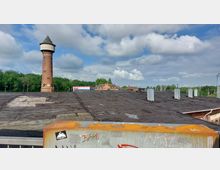 Industriegebiet mit einem hohen, roten Wasserturm im Zentrum, umgeben von Bäumen und Gebäuden im Hintergrund. Im Vordergrund sind ein großes Flachdach mit Lüftungsschächten sowie ein gelblich-graues Geländer sichtbar, das mit Graffiti bemalt ist.