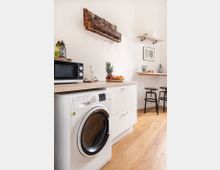 Kleine Küche mit Holzboden, weißer Waschmaschine und Arbeitsplatte aus Holz. An der Wand hängt ein rustikales Holzregal mit Weingläsern, daneben ein Regalbrett mit Dekorationsgegenständen; im Hintergrund stehen zwei schwarze Barhocker.