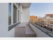 Balkon einer Wohnanlage mit weißer Fassade, großen Fenstern und einer simplen Holzbank entlang der Wand. Im Hintergrund sind Baumkronen sowie umliegende Gebäude mit roten und weißen Dächern sichtbar.