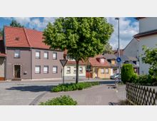 Straßenszene in einer Wohngegend mit Reihenhäusern, die rote Dächer und bunte Fassaden in Braun, Gelb und Beige haben. Im Vordergrund stehen ein Baum, ein Gehweg, ein Verkehrszeichen und ein niedriger Holzzaun; im Hintergrund sind Autos und eine Fußgängerampel zu sehen.