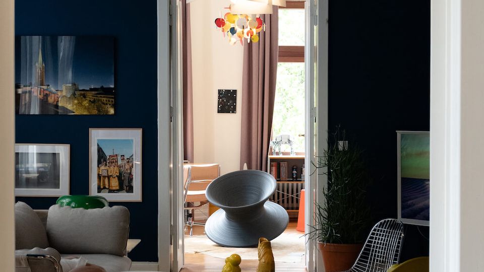 Modern living room with dark blue walls, framed artwork, and a mix of contemporary chairs around a glass coffee table on a light rug. A geometric pendant light hangs over the seating area, and open double doors lead to a brighter adjoining room with tall windows and curtains.