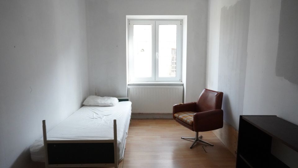 Schmales, schlichtes Schlafzimmer mit weißen Wänden und hellem Holzfußboden, einem Einzelbett an der linken Wand und einem braunen Sessel. An der Stirnwand befindet sich ein großes Fenster mit Heizkörper darunter; rechts steht ein dunkles Regal/Schrankmöbel.