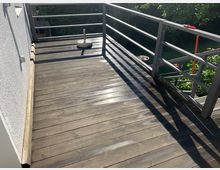 Holzbalkon mit seitlichem Metallgeländer und angrenzender verputzter Hauswand. Im Hintergrund sind grüne Bäume und ein Gartenbereich sichtbar.