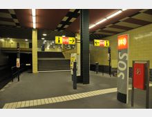 U-Bahn-Station mit gelb gekachelten Wänden, schwarzen Stahlträgern und einer Treppe, die zu einer oberen Ebene führt. Im Vordergrund befinden sich ein Fahrkartenautomat, ein SOS-Notrufterminal und ein Feuerlöscherkasten. Gelbe Hinweisschilder mit Richtungspfeilen hängen von der Decke.