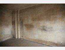 Ein leerer Raum mit verblassten, schmutzig-beigen Wänden, die Abnutzungs- und Schimmelflecken aufweisen. Der Boden ist grau, und an den Wänden sind Steckdosen sowie kleine Beschädigungen sichtbar.