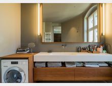 Modernes Badezimmer mit einem breiten Waschtisch aus Holz und einem weißen Waschbecken, darüber ein großer beleuchteter Spiegel. Neben dem Waschtisch befindet sich eine Waschmaschine, und auf den offenen Regalböden liegen gefaltete Handtücher; rechts sind Pflegeprodukte und Zubehör ordentlich angeordnet.