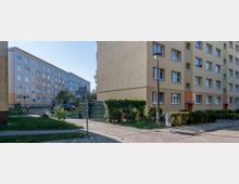 Wohnsiedlung mit mehrstöckigen Wohngebäuden in schlichten Farben (Gelb und Beige), umgeben von kleinen Grünflächen und Hecken. Im Vordergrund sind ein Gehweg, ein Laternenmast, Büsche und parkende Autos zu sehen.