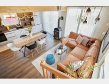 Wohnraum mit offenem Design, bestehend aus einer Küche mit weißen Schränken und Holzarbeitsplatte, einem Esstisch mit gemischten schwarzen und weißen Stühlen sowie einem angrenzenden Wohnzimmer mit cognacfarbener Eckcouch, Couchtisch, Kaminofen und großen bodentiefen Fenstern mit lichtdurchlässigen Vorhängen.