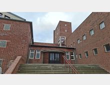 Backsteinbau mit moderner Architektur, das den Eingang eines Stadtbads zeigt. Zu sehen sind breite Treppenstufen mit einem roten Geländer, ein zentraler Eingang mit gewölbtem Vordach und hohe, schlichte Fenster.