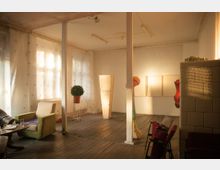 Ein hell erleuchteter Raum mit Holzfußboden, weißen Wänden und einem Vorhang aus dünnem Stoff mit dezentem Kreuzmuster. Der Raum ist mit einem grünen Sessel, einem kleinen runden Tisch, einer Topfpflanze, einer Stehleuchte und einzelnen Kunstobjekten ausgestattet.