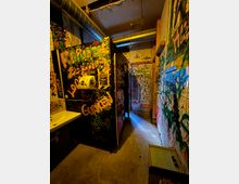 Schmaler, industriell wirkender Innenraum mit Betonboden, sichtbaren Lüftungsrohren an der Decke und warmem Kunstlicht. Wände, Türen und Trennwände sind dicht mit Graffiti und Stickern bedeckt; links ist ein kleines Waschbecken zu sehen.